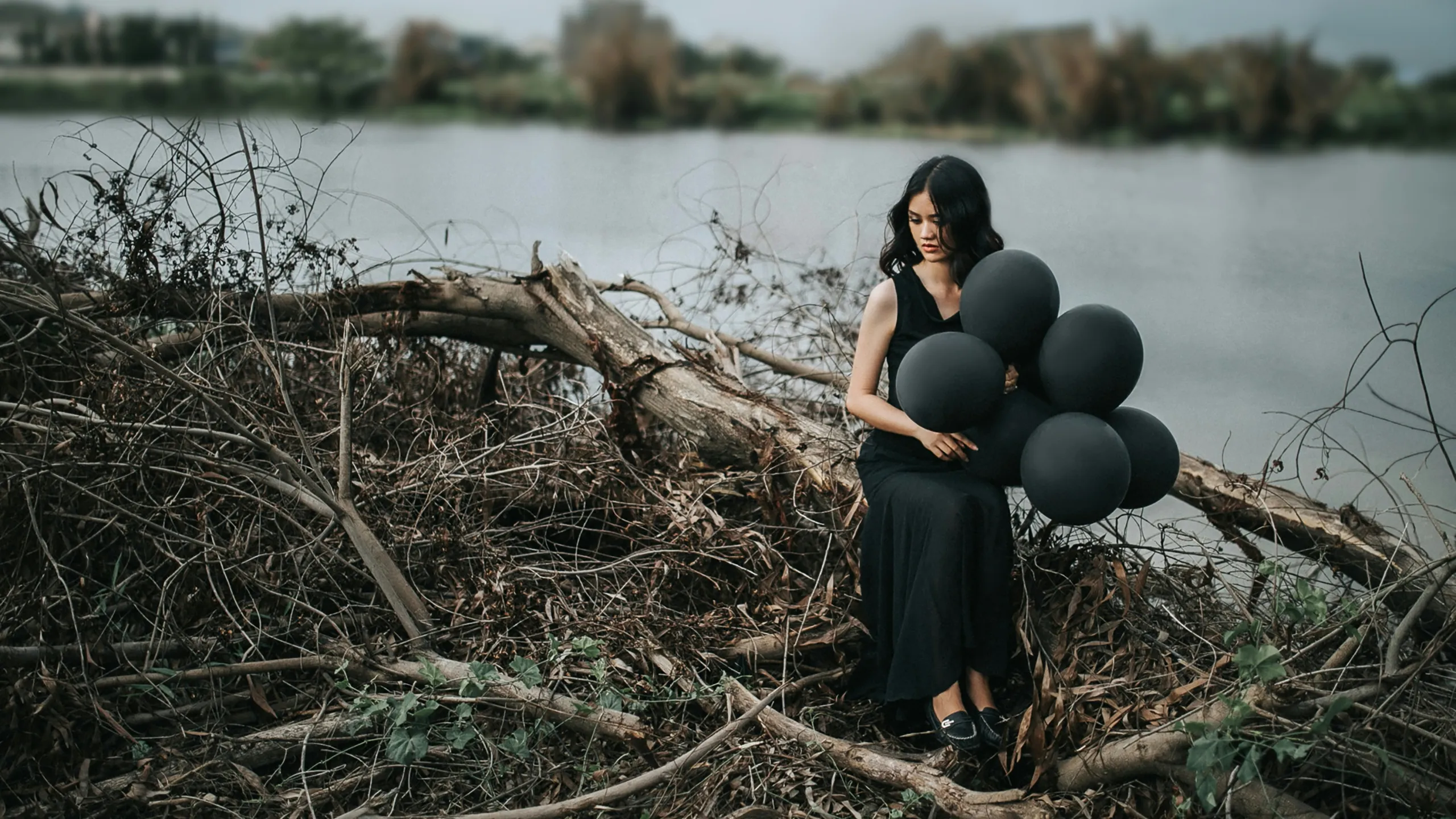 A woman in black sitting by a lake, holding dark balloons